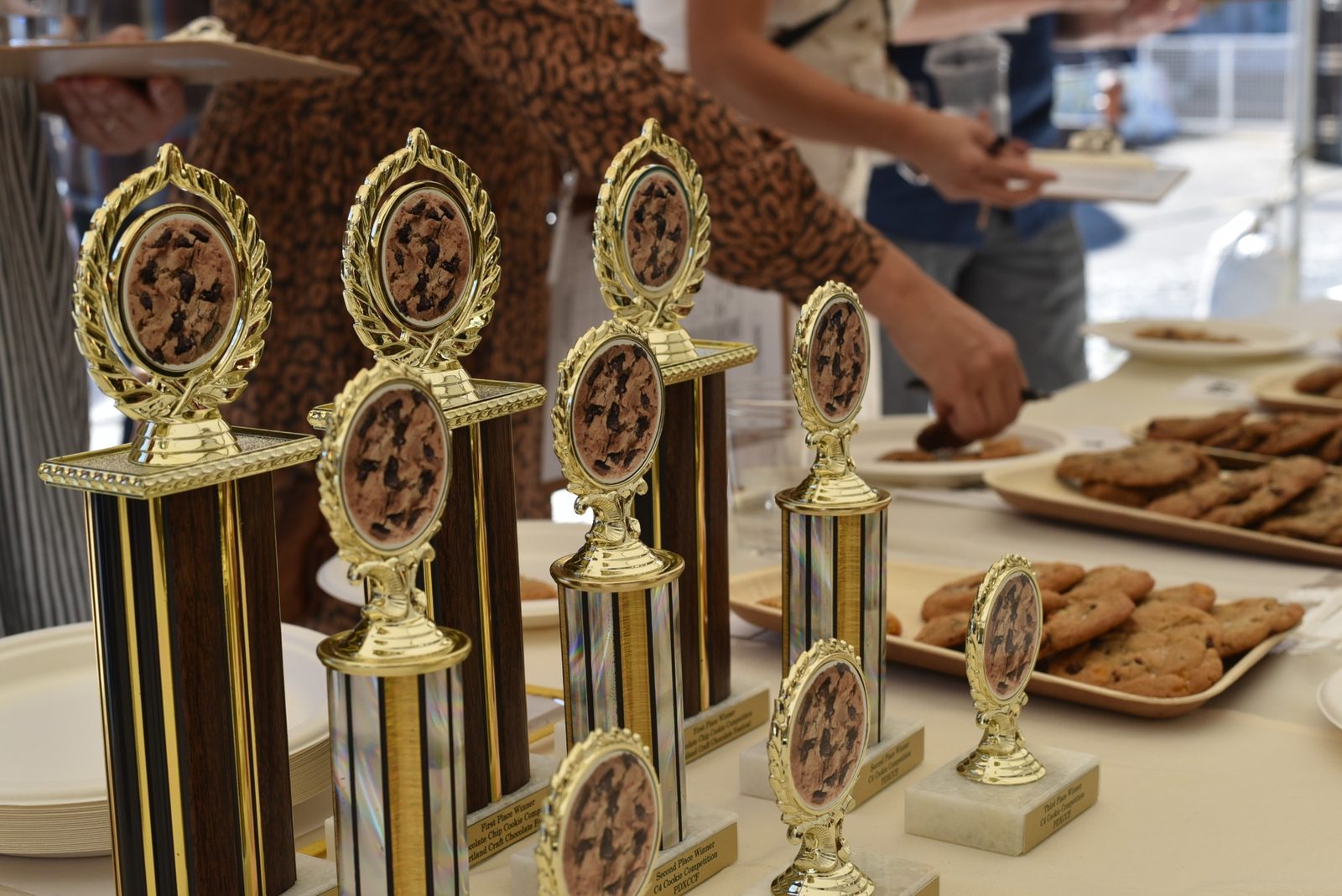 First-, second- and third-place C4 trophies, each topped with a chocolate chip cookie, on the judging table with trays of cookies and judges' clipboards behind them.